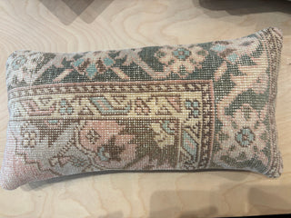 A rectangular needlepoint pillow with a vintage-style floral and geometric pattern in muted shades of green, beige, pink, and blue, displayed on a light wood grain surface.