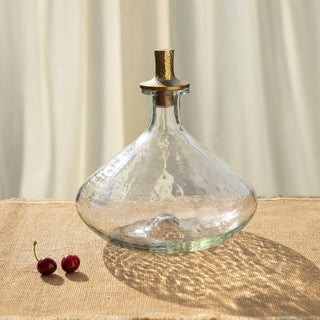 A clear glass decanter with a wide bottom and textured surface and a bronze-colored hammered aluminum stopper