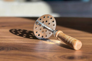 A stainless steel potato masher with a perforated head and acacia wood handle, placed on a wooden surface.