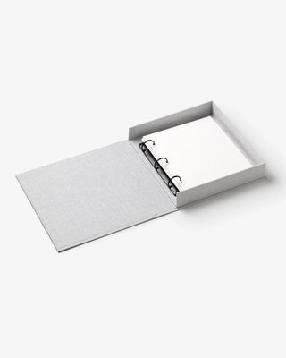 A light gray fabric-covered ring binder notebook shown open on a clean white background, with white pages visible inside and black metal rings.