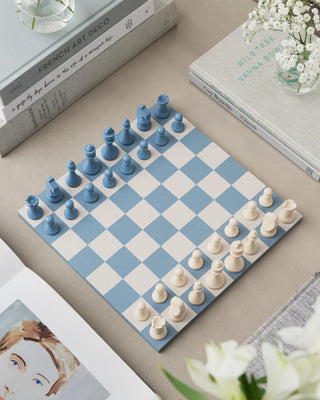 Chessboard with blue and white pieces on a table with books and flowers.