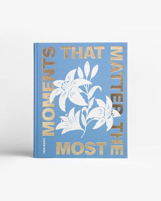 A hardcover art book titled 'Moments That Matter Most' by Port & Company, featuring a light blue cover with white lily illustrations and metallic gold lettering, set against a clean white background.