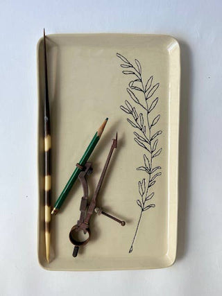 A rectangular beige ceramic tray with rounded corners, featuring a black botanical line drawing of a leafy sprig on the right side. The tray is styled with vintage writing and drawing tools, including a tortoiseshell pen, a green pencil, and a metal compass, all set against a plain white background.