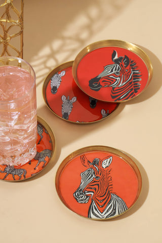 A set of round coasters with gold-toned rims, featuring a bold zebra design on a bright red and orange background. One coaster is shown holding a clear glass filled with pink liquid, all arranged on a warm beige surface with soft shadows.