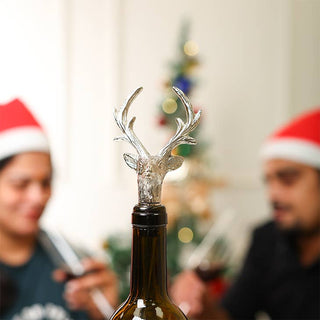 A silver reindeer head bottle stopper on a dark glass wine bottle, with two blurred people wearing Santa hats in the background near a Christmas tree.
