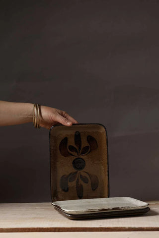 A hand wearing gold bangles holds a square brown ceramic tray with a black floral pattern, with a second tray stacked on a wooden surface in front, against a dark brown background.