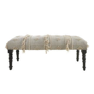 An upholstered bench with a textured cream woven fabric seat featuring fringe and tufted details, supported by four dark turned wooden legs, shown against a plain white background.