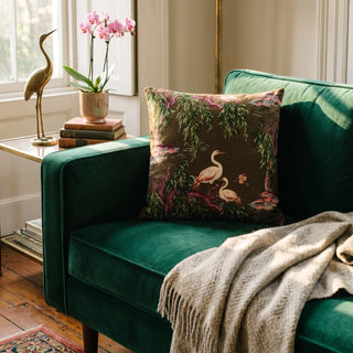 A decorative throw pillow with a brown background and a heron and bird pattern, styled on a deep green velvet armchair next to a beige knit throw blanket.