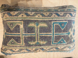A square woven throw pillow with a geometric pattern in blue, beige, and gray, resting on a light wood surface.