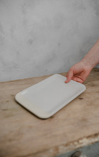 A hand holding a rectangular white ceramic tray with rounded corners on a rustic wooden table against a textured gray wall.