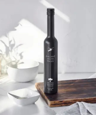 A tall black bottle of Ancient Olive Trees extra virgin olive oil stands on a wooden cutting board on a light marble surface, with white bowls and soft natural shadows in the background.