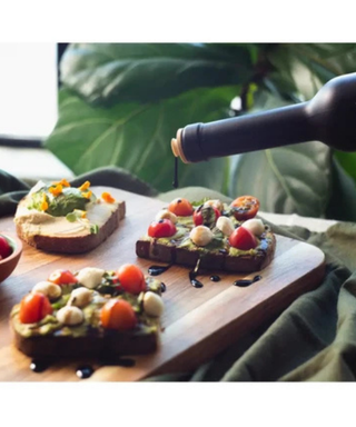 Three slices of toasted bread topped with avocado, cherry tomatoes, mozzarella balls, and herbs are arranged on a wooden board, with a dark bottle pouring dressing over the food. Large green leaves and a draped fabric are visible in the softly lit background.