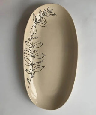 An oval ceramic serving platter in a warm beige color, decorated with a minimalist black line drawing of a leafy branch running down the left side, set against a neutral gray background.