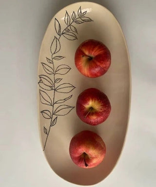 An oval beige ceramic serving platter with a black line-art botanical leaf pattern, containing three red apples arranged vertically on a light gray background.