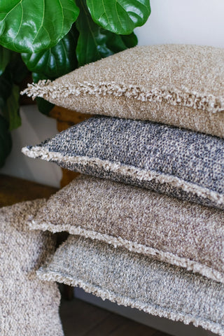 A stack of textured throw pillows in neutral tones, including beige, gray, taupe, and light gray, with fringed edges, styled indoors next to a green houseplant.
