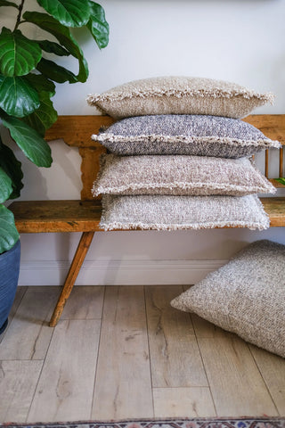 Four textured woven throw pillows in neutral beige, gray, and cream tones are stacked on a rustic wooden bench, with a matching pillow resting on the light wood floor. A large green potted plant is visible on the left, and a white wall forms the background.