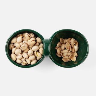 A top-down view of a dark green ceramic double-compartment snack dish filled with pistachios on a light gray background.