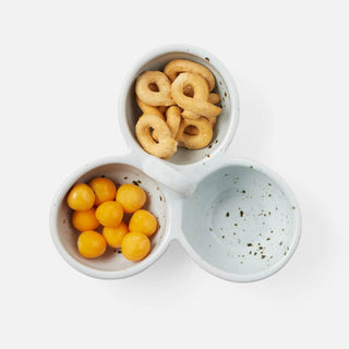A three-compartment ceramic snack plate in a speckled light blue-gray glaze, shown from a top-down view on a white background. One compartment holds golden ring-shaped crackers, another holds small round orange candies, and the third is empty.