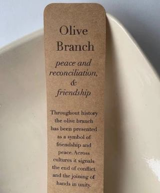 A rectangular olive branch-shaped bookmark or card made of natural kraft paper with printed black text, leaning against a cream-colored ceramic dish.