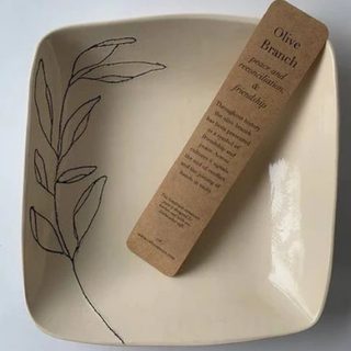 A square cream-colored ceramic plate with a black line-art olive branch design on the left side, featuring a brown kraft paper bookmark titled 'Olive Branch' placed diagonally on top.