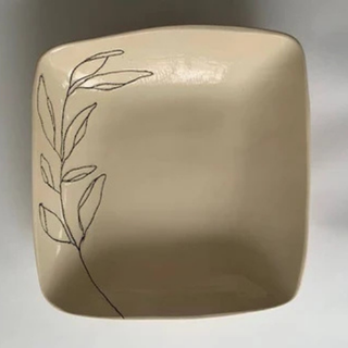 A square ceramic plate with rounded corners in a warm beige color, featuring a simple black line art illustration of a leafy branch on the left side, set against a neutral gray background.