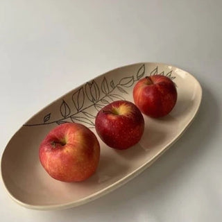 An oval beige ceramic plate with a black leaf pattern, holding three red apples on a light gray background.