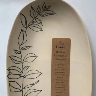 An oval ceramic sign or plaque with a cream background, featuring a black line-art leaf design along the left side and a brown rectangular label in the center that reads 'Bay Laurel' and includes a short quote. The item is photographed against a plain light background.