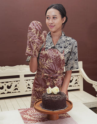 Woman in a patterned apron holding a chocolate cake with white flowers on a wooden stand.