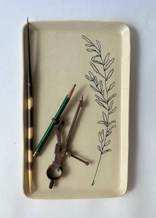 A rectangular beige ceramic tray with rounded corners, featuring a black botanical line drawing on the right side. The tray is styled with vintage drawing tools, including a green pencil, a metal compass, and a striped wooden ruler, all laid out on a light gray background.