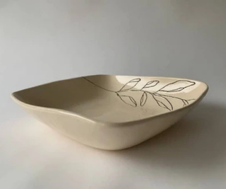 A shallow, wide ceramic bowl with a cream-colored glaze and a delicate hand-painted line drawing of leaves on the inside, set against a neutral light gray background.