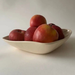 A shallow cream-colored ceramic bowl with a leaf-like outline and subtle dark line detailing, filled with red apples, set against a plain light gray background.