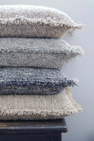 Four textured woven throw pillows in neutral tones stacked vertically on a dark wooden surface against a soft gray background.