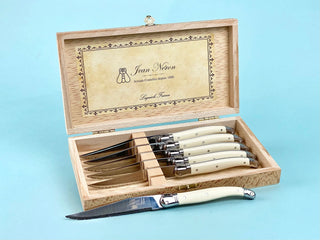 A set of Laguiole brand steak knives with ivory-colored handles, displayed in a wooden presentation box with the name 'Jean Nico' inscribed on the interior lid.